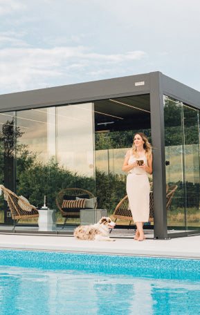 Pergolux Pergola mit Glaswänden am Pool, davor eine Frau mit Tasse und Hund an einem sonnigen Tag.