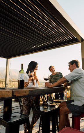 Pergolux Pergola bei Sonnenuntergang – drei Personen trinken Wein und unterhalten sich.