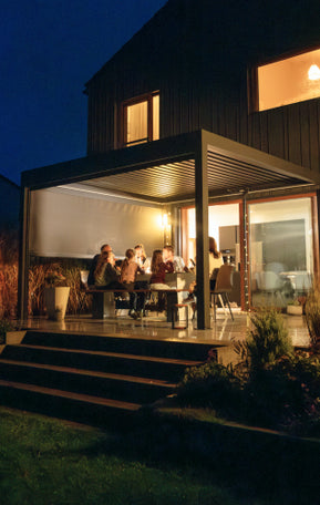 Beleuchtete Pergolux Pergola am Abend mit Personen, die gemeinsam unter dem Lamellendach sitzen und den Abend genießen.