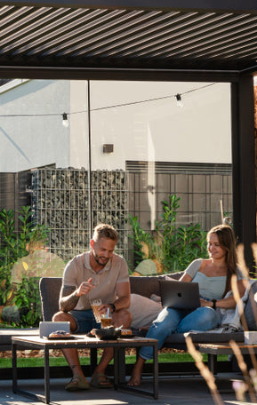 Zwei Personen sitzen unter einer Pergolux Pergola, arbeiten am Laptop und genießen den sonnigen Tag.