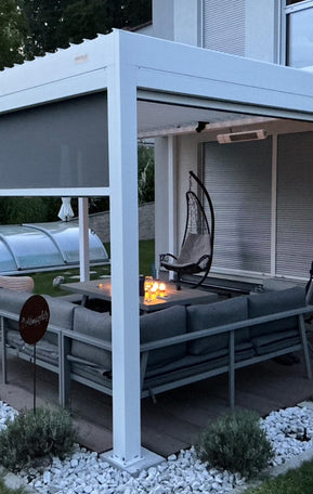 Weiße Pergolux Pergola mit heruntergelassenem Screen-Rollo und beleuchteter Lounge mit Feuerstelle bei Dämmerung.