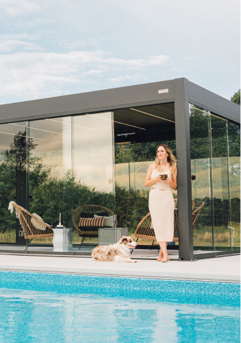 Pergolux Pergola mit Glaswänden am Pool, davor eine Frau mit Tasse und Hund an einem sonnigen Tag.