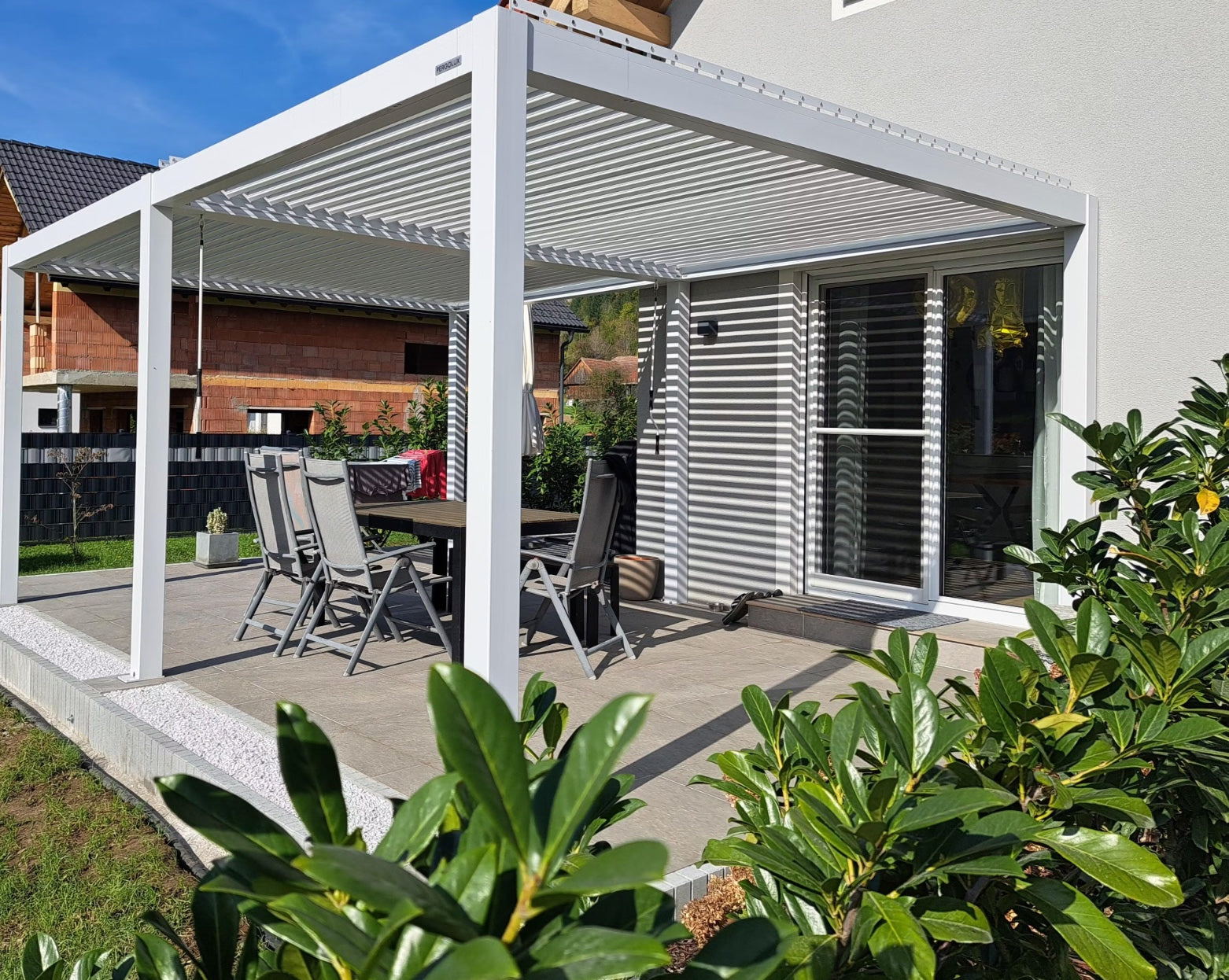 Weiße Pergolux Pergola auf einer Terrasse mit Esstisch und Gartenstühlen im Sonnenschein.